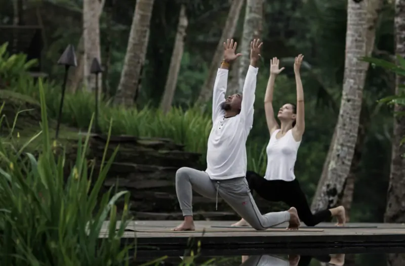 Man and woman performing yoga poses on a wooden platform amid lush palm trees and greenery in Bali.