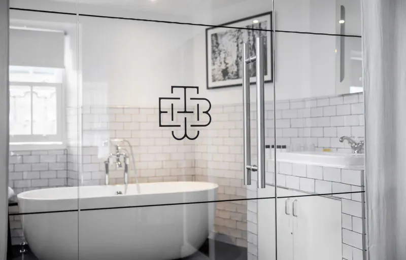 Glass shower door with 'TBB' logo in elegant white bathroom featuring freestanding tub, subway tiles, and window.