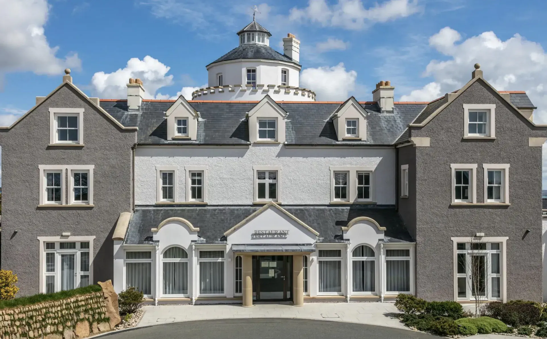 Twr y Felin hotel, a grand gray stone building with white rotunda tower and entrance, set in landscaped gardens under partly cloudy sky.