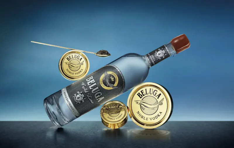 Beluga Gold Line vodka bottle tilted on dark surface with gold caviar tins and spoon on blue gradient background.
