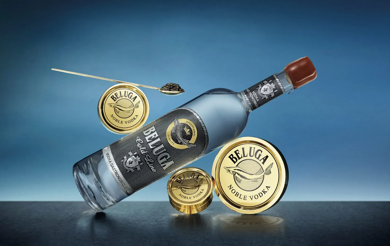 Beluga Gold Line vodka bottle tilted on dark surface with gold caviar tins and spoon on blue gradient background.
