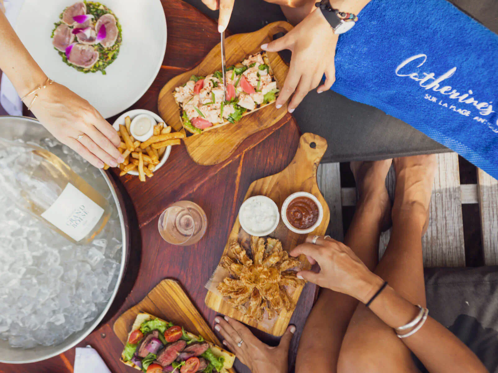 Top-down view of hands sharing Catherine's Café seafood platters, fries, and rosé on wooden boards by the beach.
