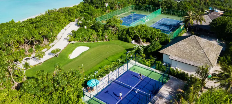 Aerial view of tropical resort with blue tennis courts, players in action, pool, beach, and lush greenery.