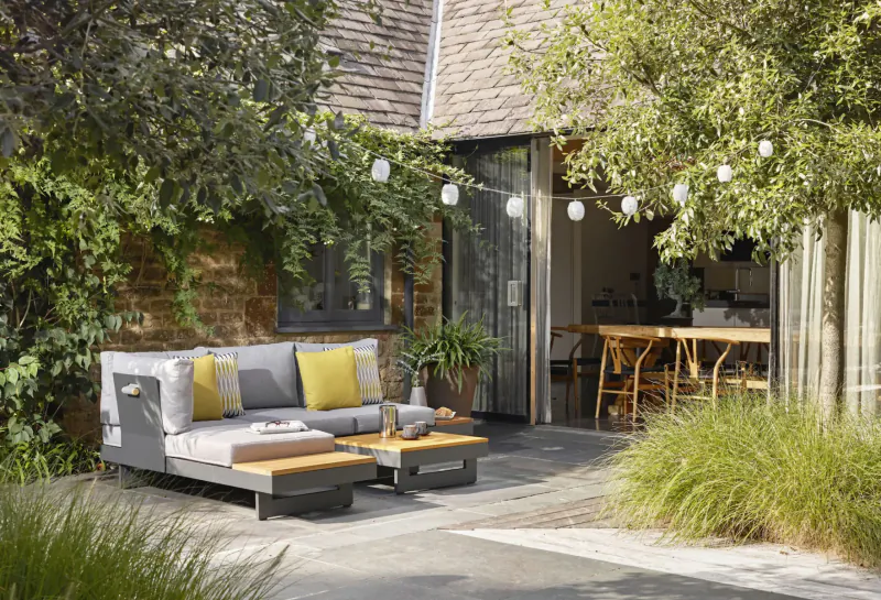 Modern grey Bramblecrest garden sofa with yellow cushions and coffee table on lush patio near open kitchen doors