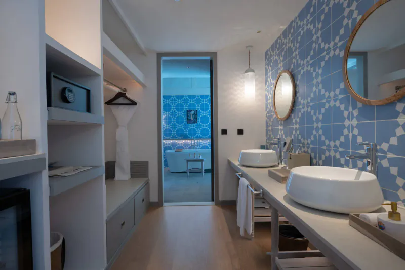 Luxurious hotel bathroom with blue patterned tile wall, double sinks, and open door to bedroom suite