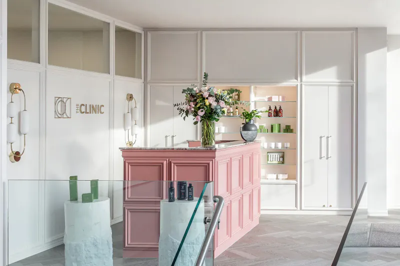 Stylish Clinic Holland Park reception with pink reception desk, flowers, skincare products on shelves, and 'CLINIC' logo.