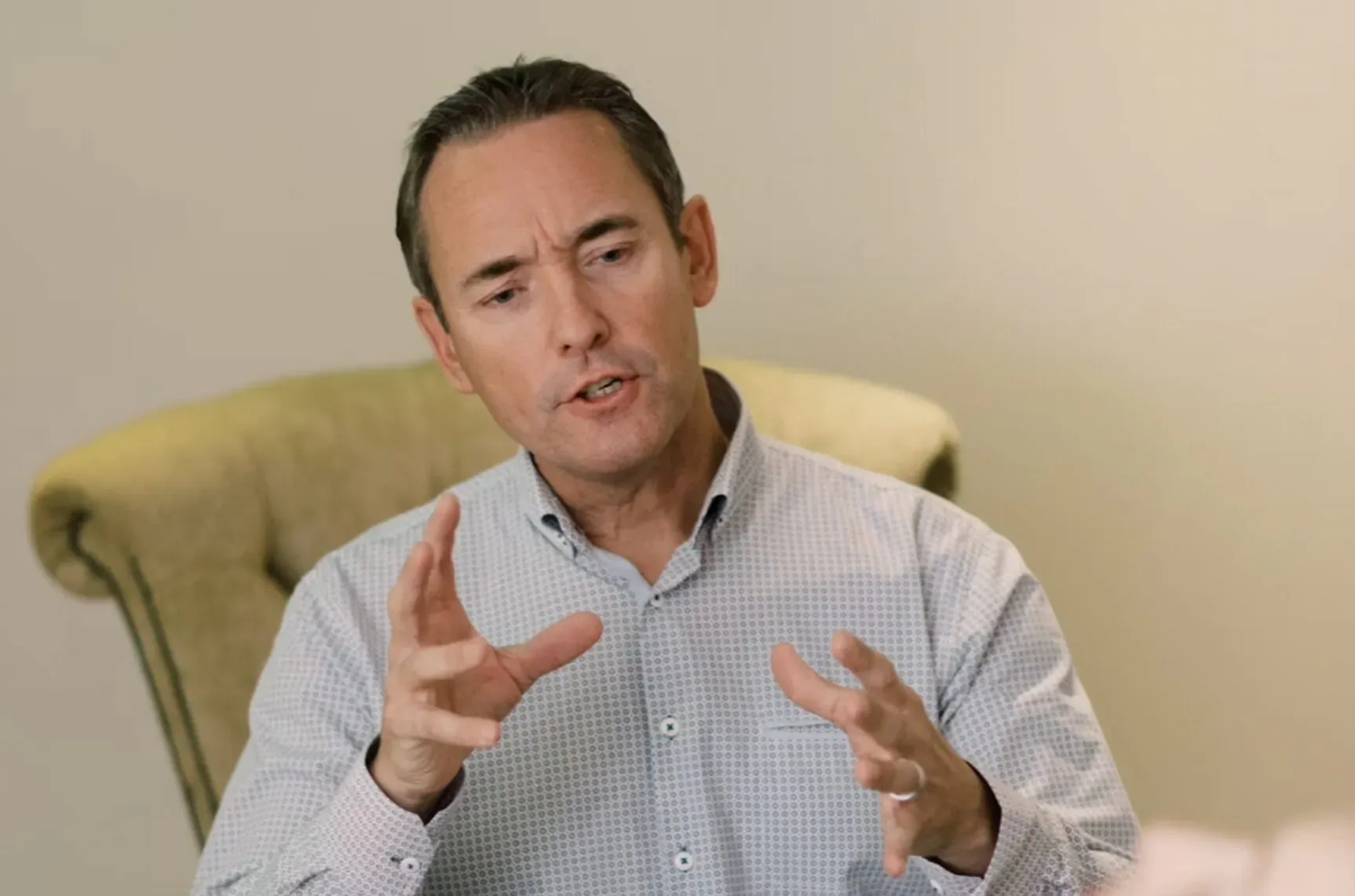Man in light blue shirt gesturing with hands while seated in green armchair, speaking in interview setting.