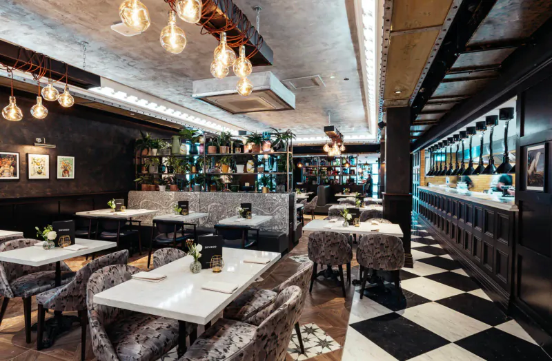 Six by Nico Canary Wharf restaurant interior with Ancient Rome theme, black-and-white checkered floor, velvet chairs, plants, and bar.