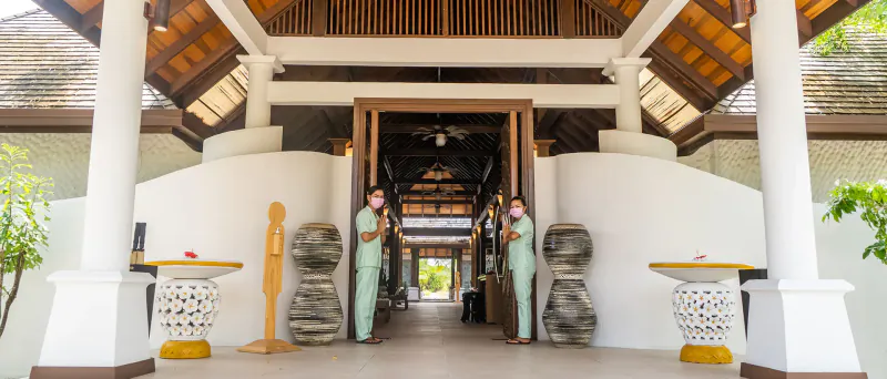 Hideaway Beach Resort entrance: two women in green uniforms flank wooden statues beside curved white pillars and tropical plants.