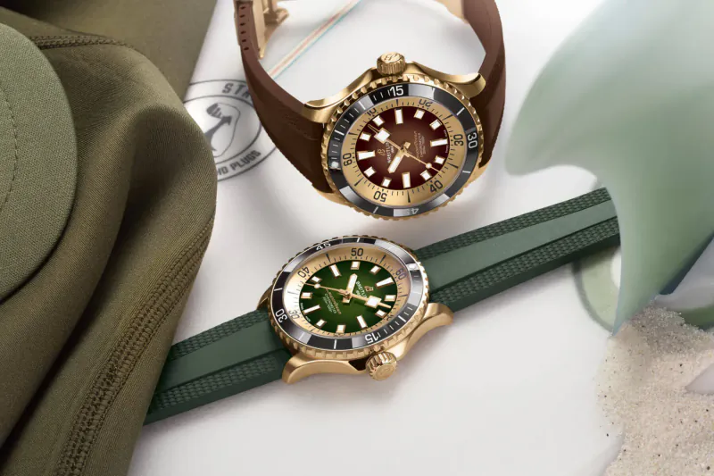 Two Breitling Superocean watches on green and brown straps beside a military-style hat on white surface.