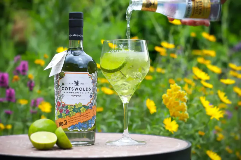 Pouring Cotswolds Wildflower Gin No. 3 into a lime gin and tonic glass, with lime wedges, on table amid wildflowers.