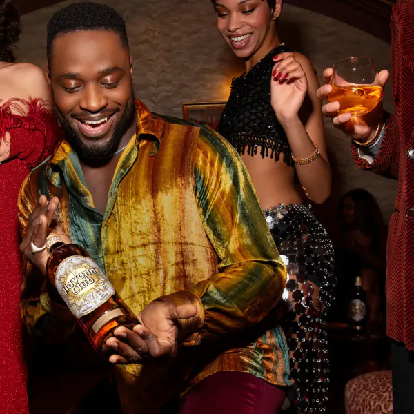 Smiling man in colorful shirt holds limited-edition Havana Club x Jonathan Raksha bottle, dancing with stylish women in gallery.