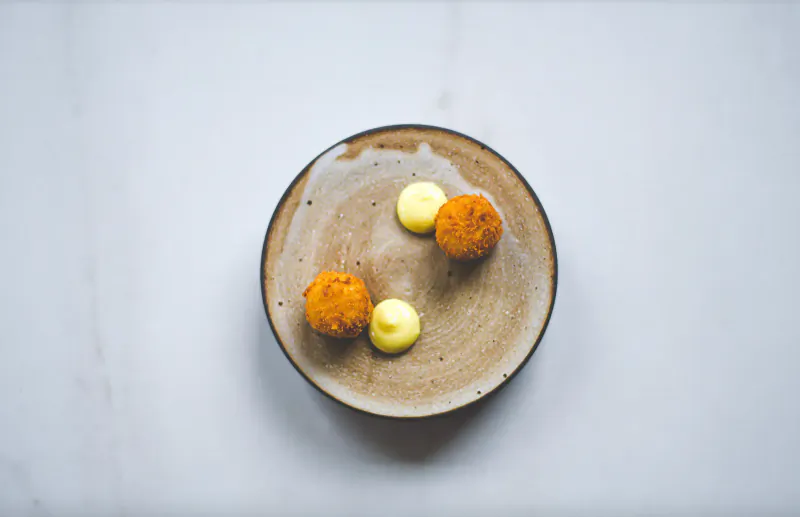 Top-down view of two golden croquettes and yellow sauce dollop on a textured brown plate on white surface