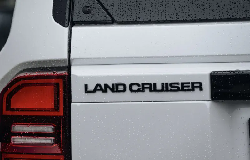 Close-up of white Toyota Land Cruiser rear side with 'LAND CRUISER' badge, red taillight, and raindrops