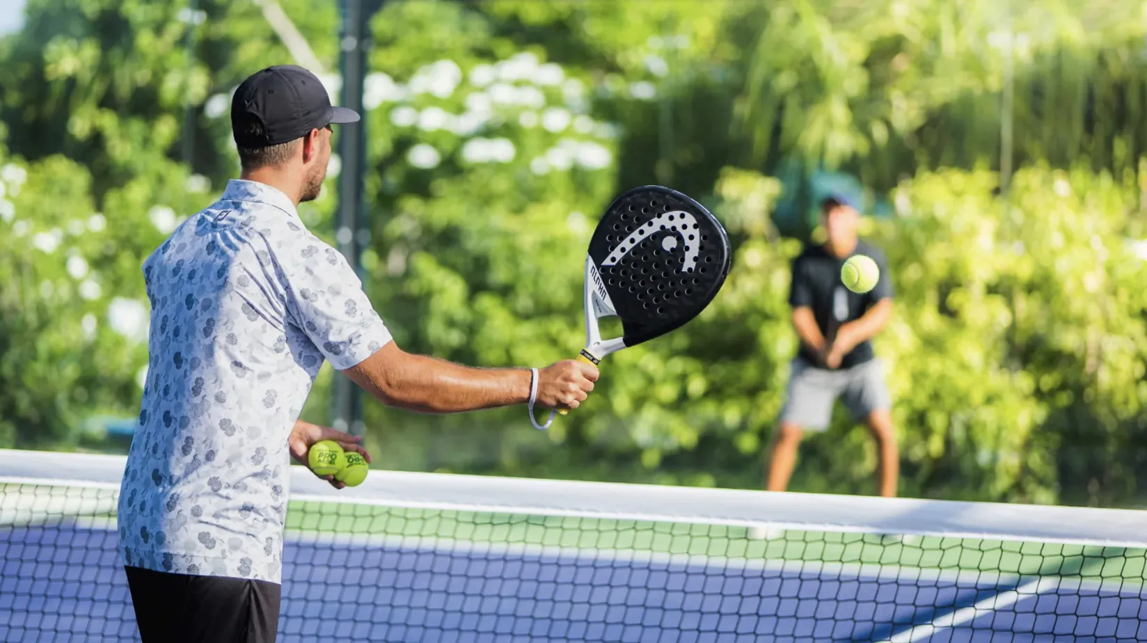 Two men with Head padel rackets ready to play padel tennis on outdoor blue court at Hideaway Beach Resort, lush greenery backdrop.
