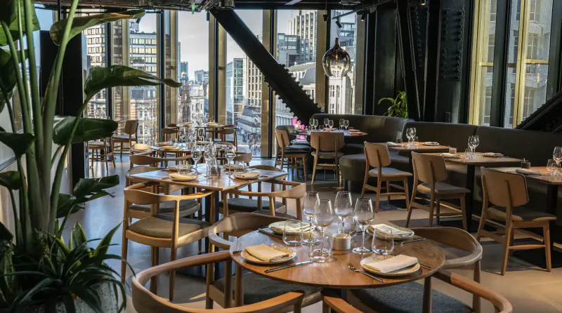 Cavo rooftop Mediterranean restaurant in Central London: elegant wooden tables set with glassware, plants, city skyline views.