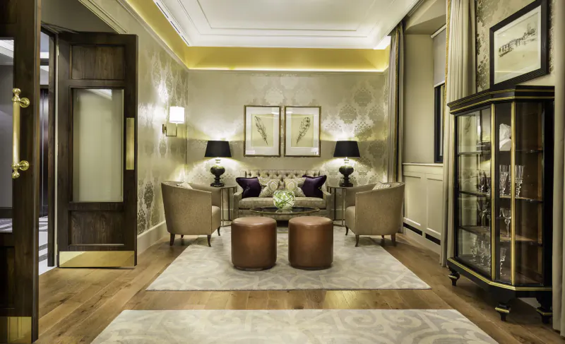 Elegant hotel lounge at Taj 51 Buckingham Gate with beige armchairs, purple cushions, copper ottomans, lamps, and display cabinet on patterned rug.