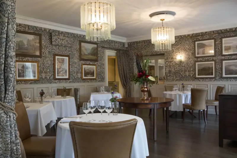 Elegant dining room at Rothay Manor with round tables set for meals, ornate wallpaper, chandelier lights, and floral arrangements.