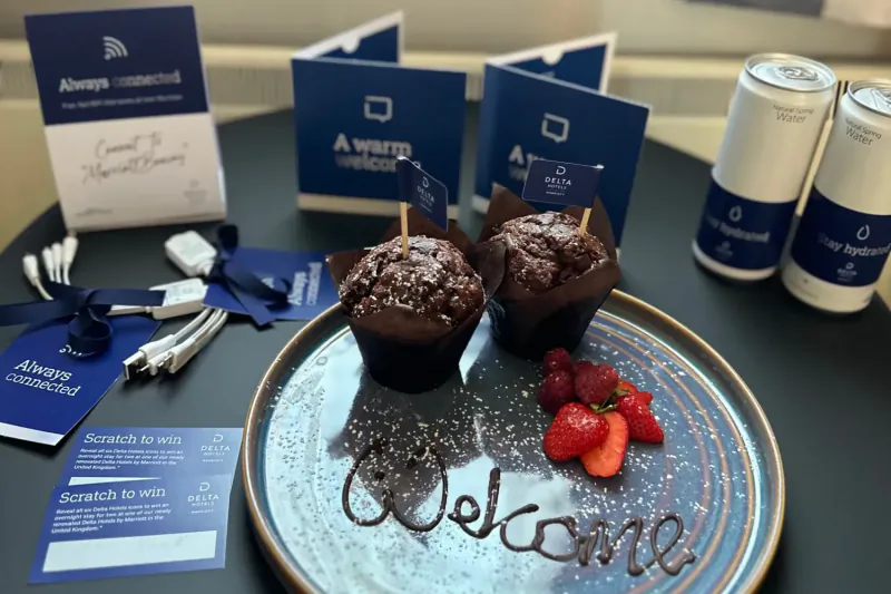 Two chocolate muffins with raspberries on a plate with 'Welcome' written in sauce at Delta Hotel Swansea, plus branded cards and cans.