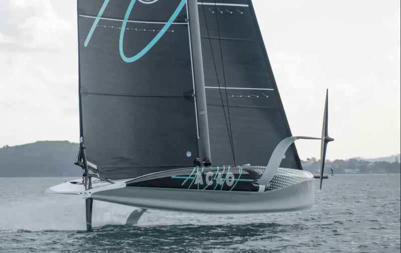 AC40 foiling yacht 'Ancasta' with cyan '10' on sail and 'AC40' on hull, speeding over water near hills.
