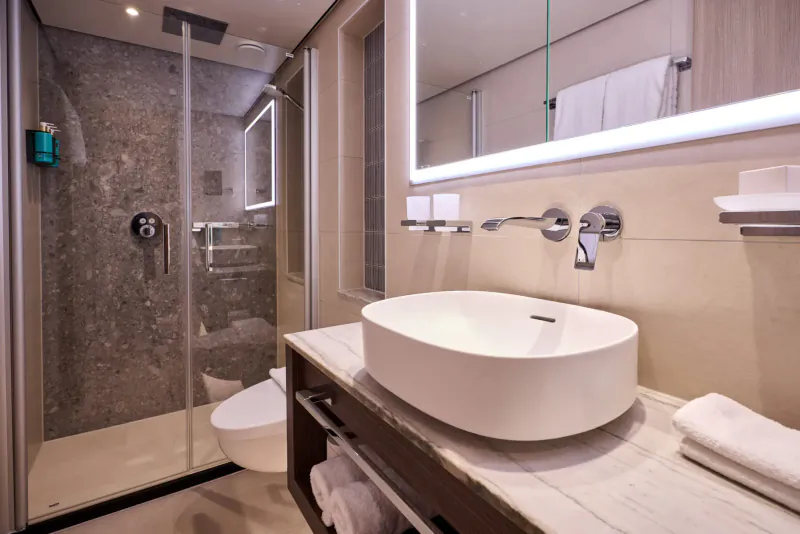 Modern luxury cruise ship bathroom with glass shower, white vessel sink, backlit mirror, and towels.