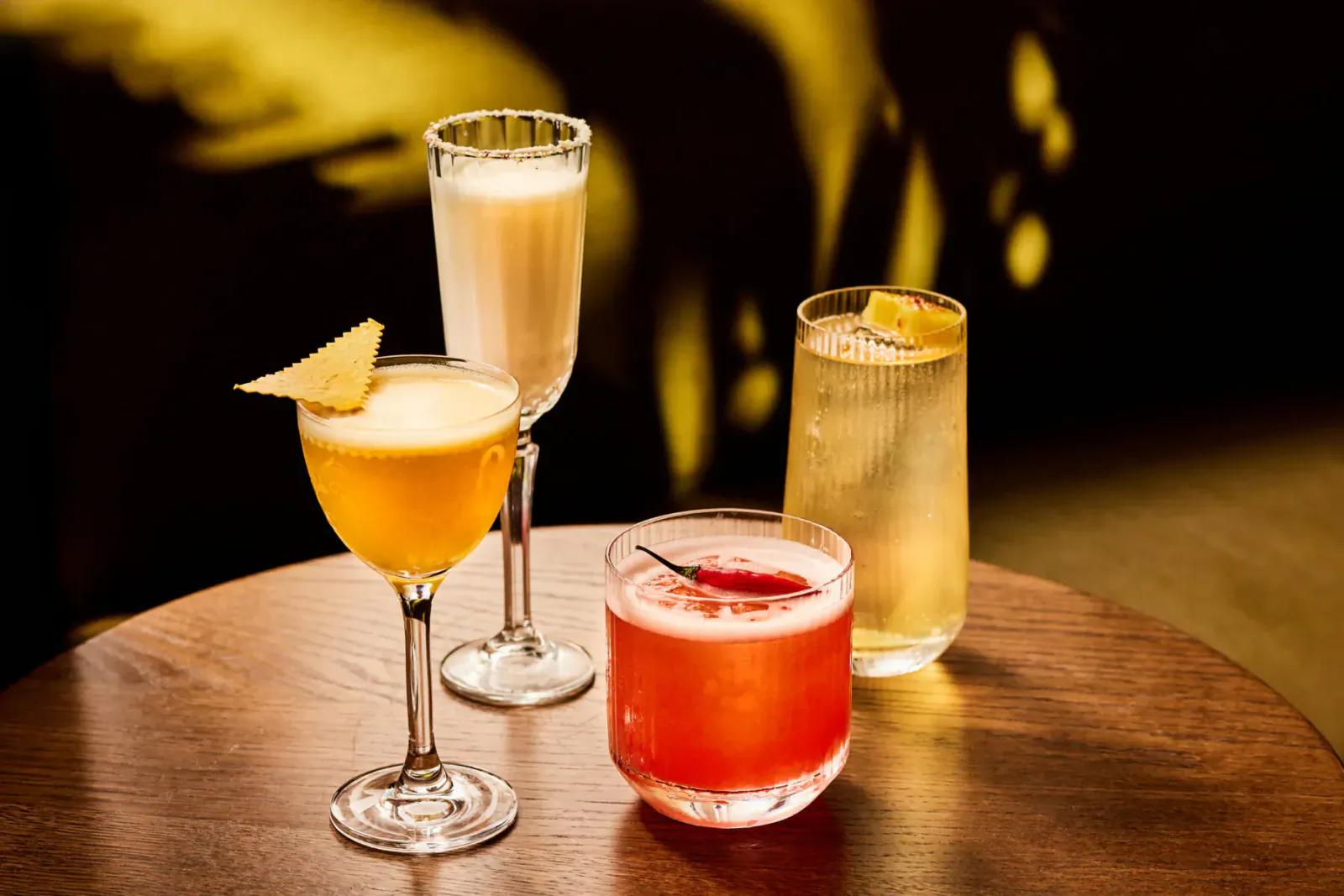 Four colorful cocktails on a wooden table: yellow with lemon, beige champagne, red chili, and pale gimlet, warm lighting.