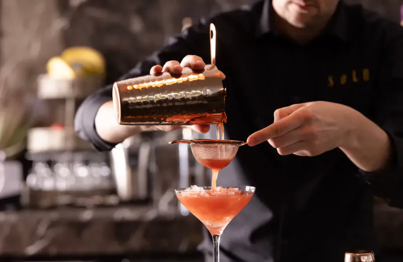 Bartender in black uniform straining pink cocktail from gold shaker into martini glass at Solo restaurant bar