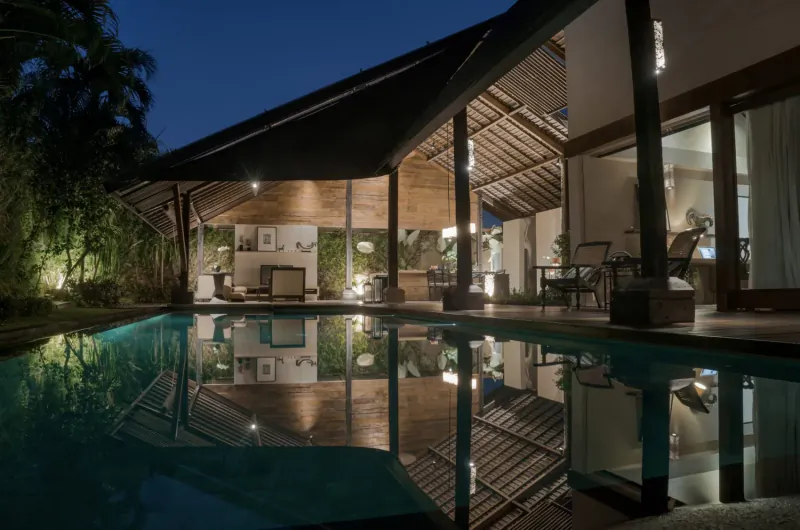Twilight view of Ametis Villa Bali: modern wooden pavilion, lounge seating, lush plants, and glowing infinity pool reflecting lights.