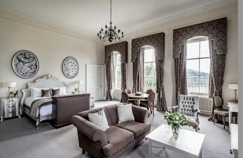 Elegant suite at Bailbrook House Hotel with king bed, brown sofa, chandelier, large windows, and hill views.
