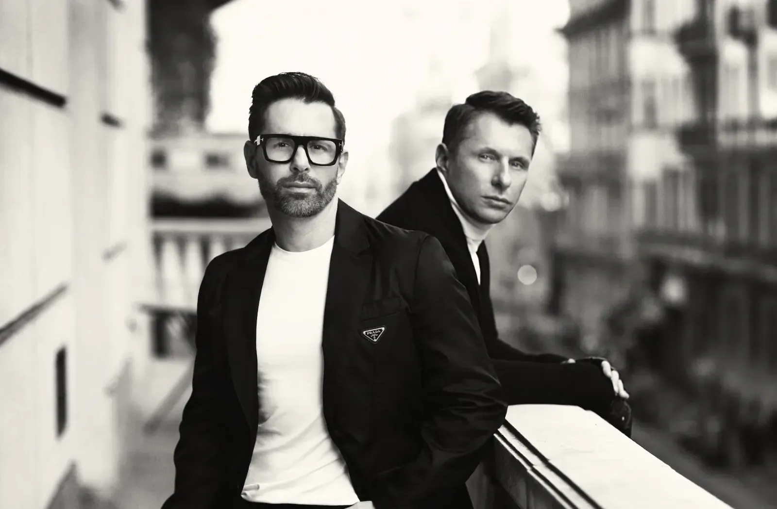 Two stylish men in suits leaning on balcony railing, black and white photo outside elegant building.