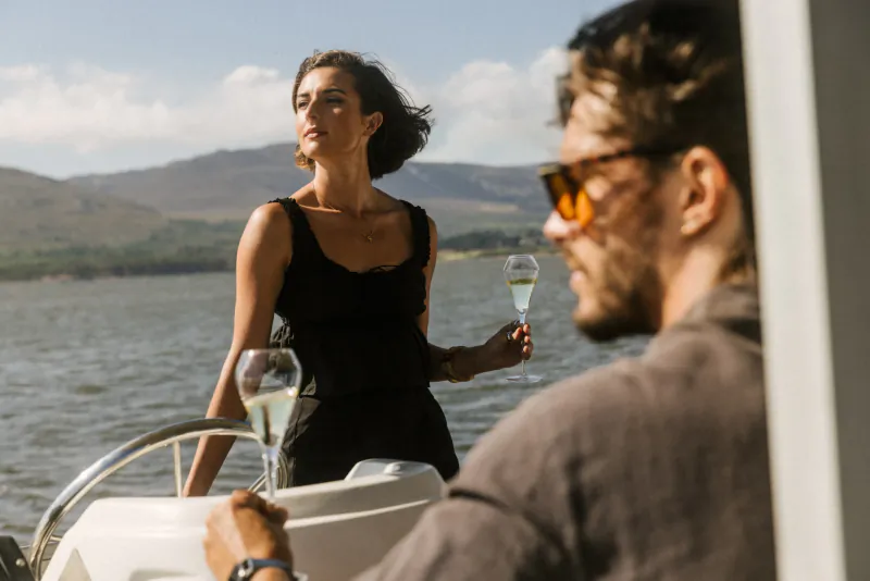 Couple on yacht at Benguela Cove, woman in black dress and man in shades holding champagne glasses, lake and mountains backdrop