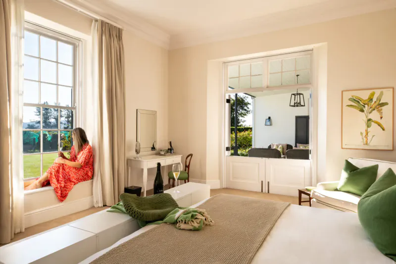 Woman in red dress sits on windowsill in elegant bedroom at Brookdale Wine Estate, gazing at green lawn view.