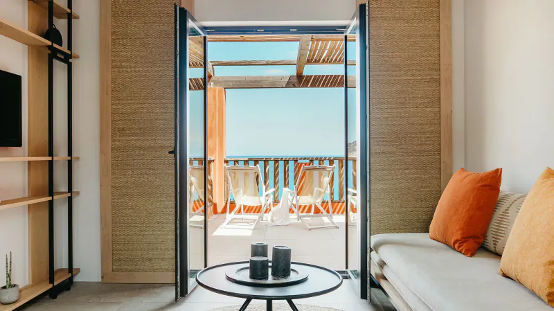 Bright hotel room with open doors to balcony overlooking sea, white sofa, orange pillows, candles on table