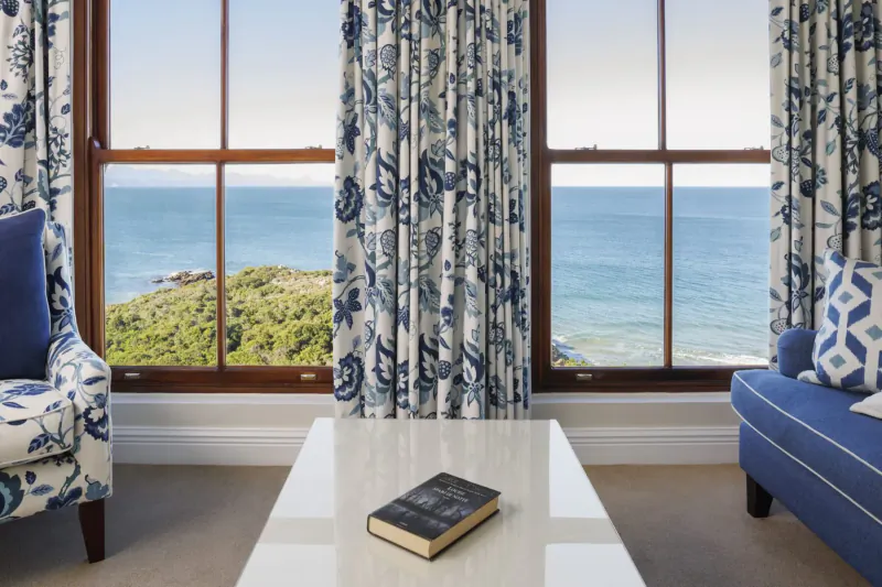 Elegant room at The Plettenberg with blue armchairs, open book on white coffee table, and ocean view through large curtained windows.