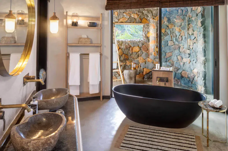 Luxurious eco-lodge bathroom with black freestanding tub, stone sinks, towels, and stone wall views of Mashatu Euphorbia camp.