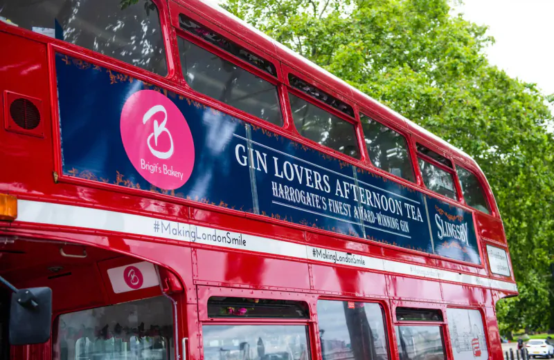 Red double-decker bus advertising Brigit's Bakery Gin Lovers Afternoon Tea, #London, trees in background