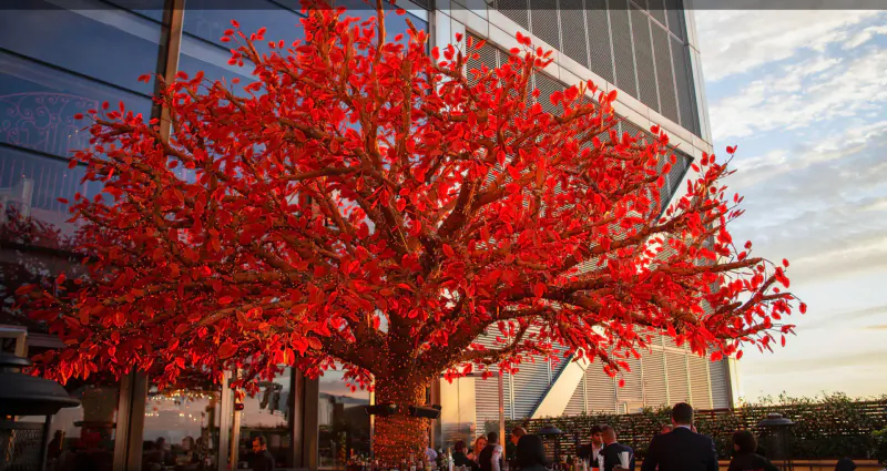 Vibrant red artificial tree beside modern glass building at SushiSamba gallery, sunset sky, blurred patrons nearby