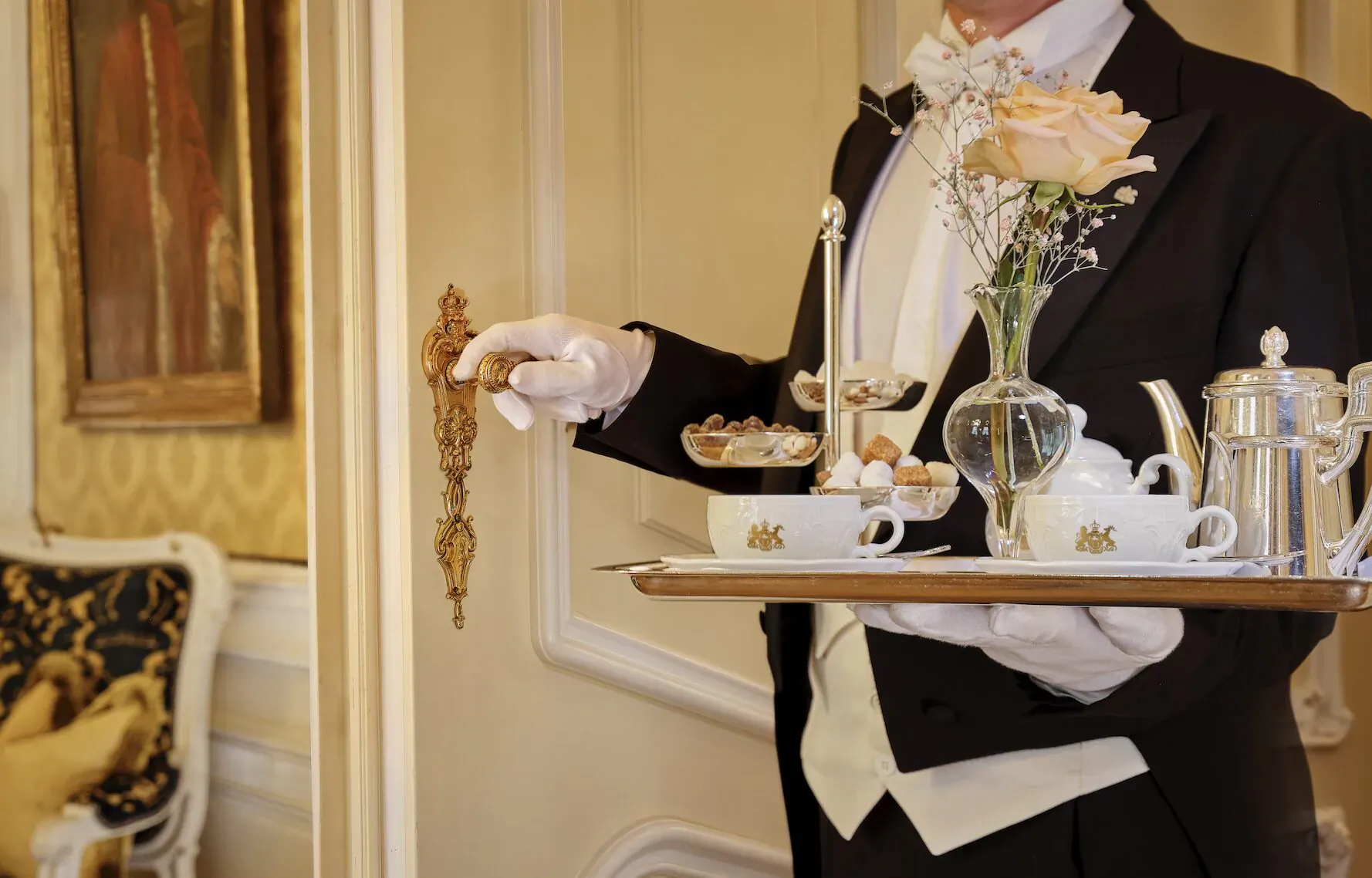 Tuxedoed waiter in white gloves holding silver tray with teapot, cups, and pastries in luxurious Hotel Imperial Vienna hotel room