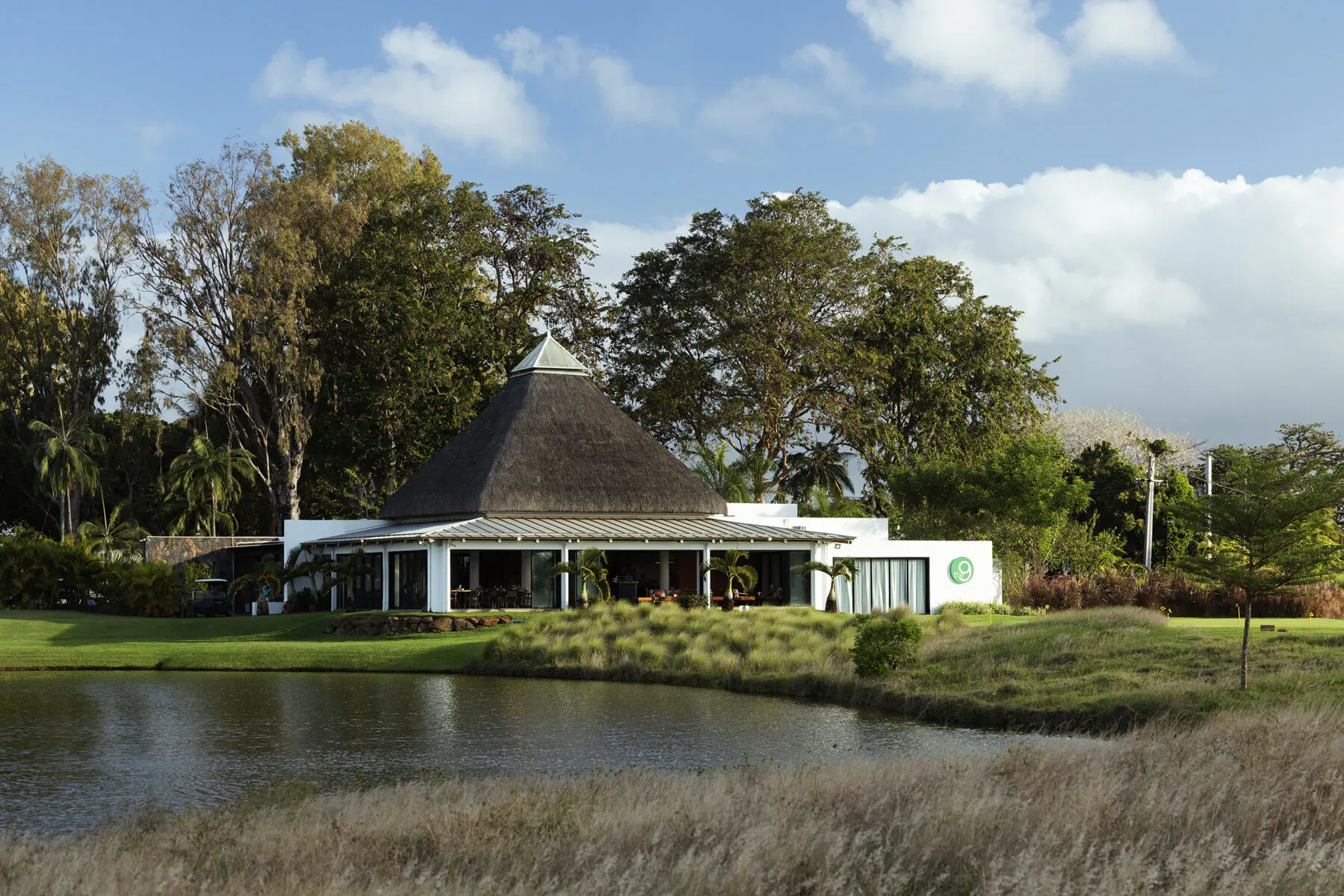 White thatched-roof clubhouse surrounded by trees and pond at Mauritius golf course under partly cloudy sky