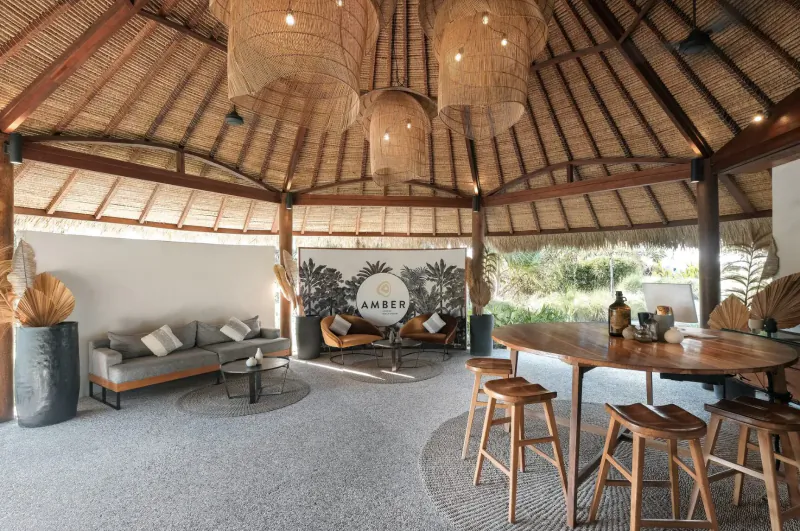 Spacious open-air dining pavilion at Amber Lombok with thatched roof, hanging lanterns, wooden table, stools, couches, and ocean views.