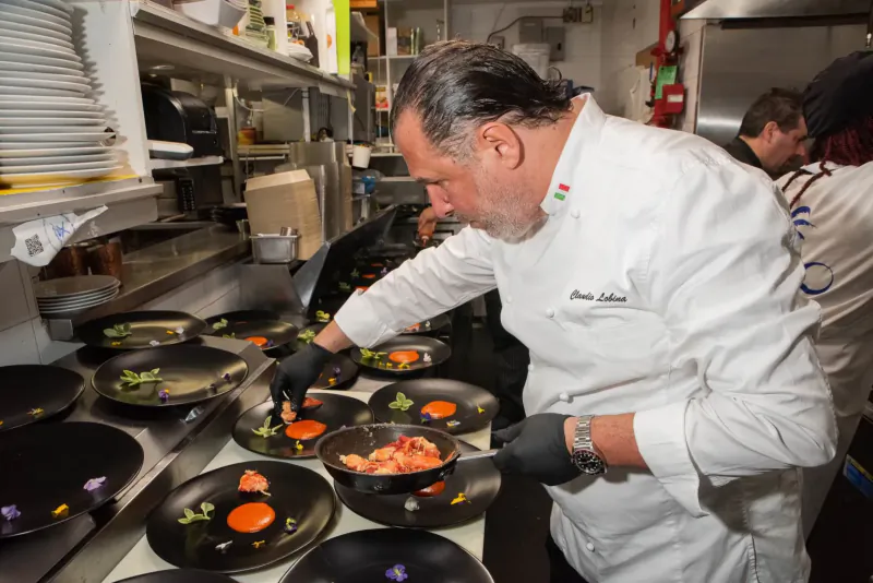 Chef Pat Martin in white uniform and gloves plating tomatoes and garnishes on black dishes in busy kitchen