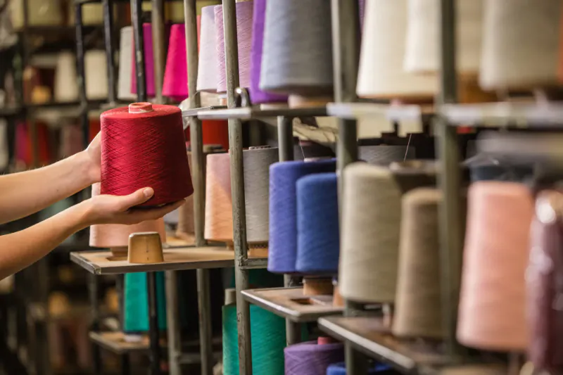 Hands holding a red yarn spool from a rack of colorful yarns in a textile factory