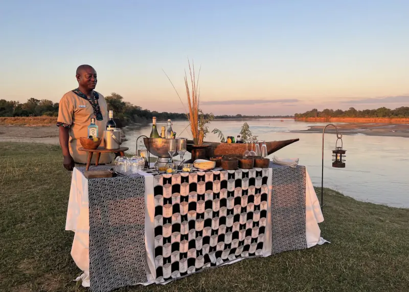African man stands by checkered tablecloth buffet with drinks, bowls, and lanterns on riverbank at sunset