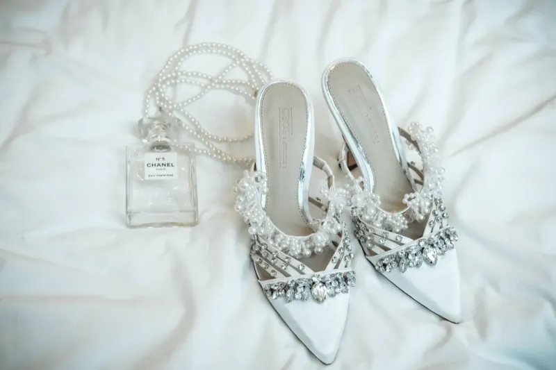 Pair of white pearl-embellished high heels with Chanel No. 5 perfume bottle on white bedding.