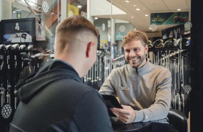 Smiling golf store associate shows tablet to customer during custom club fitting session amid golf clubs.