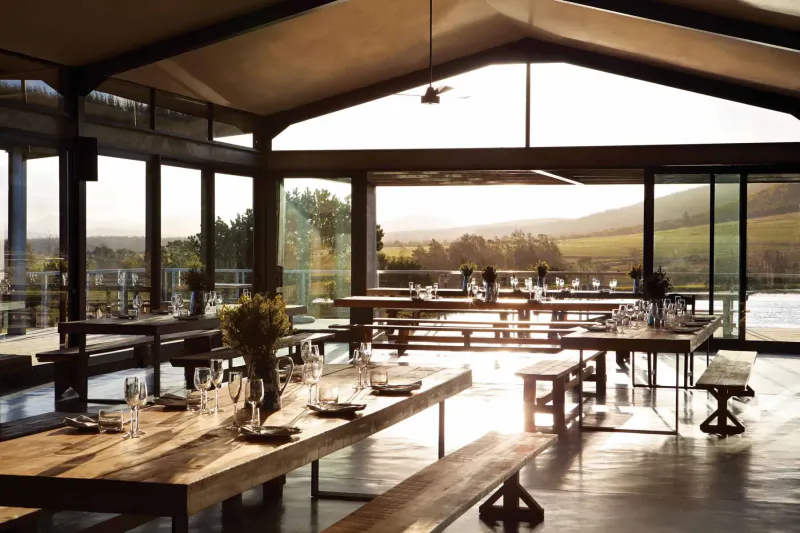 Spacious modern barn dining hall at Kay & Monty Vineyards with wooden tables set for meals, golden sunlight, vineyard and mountain views.