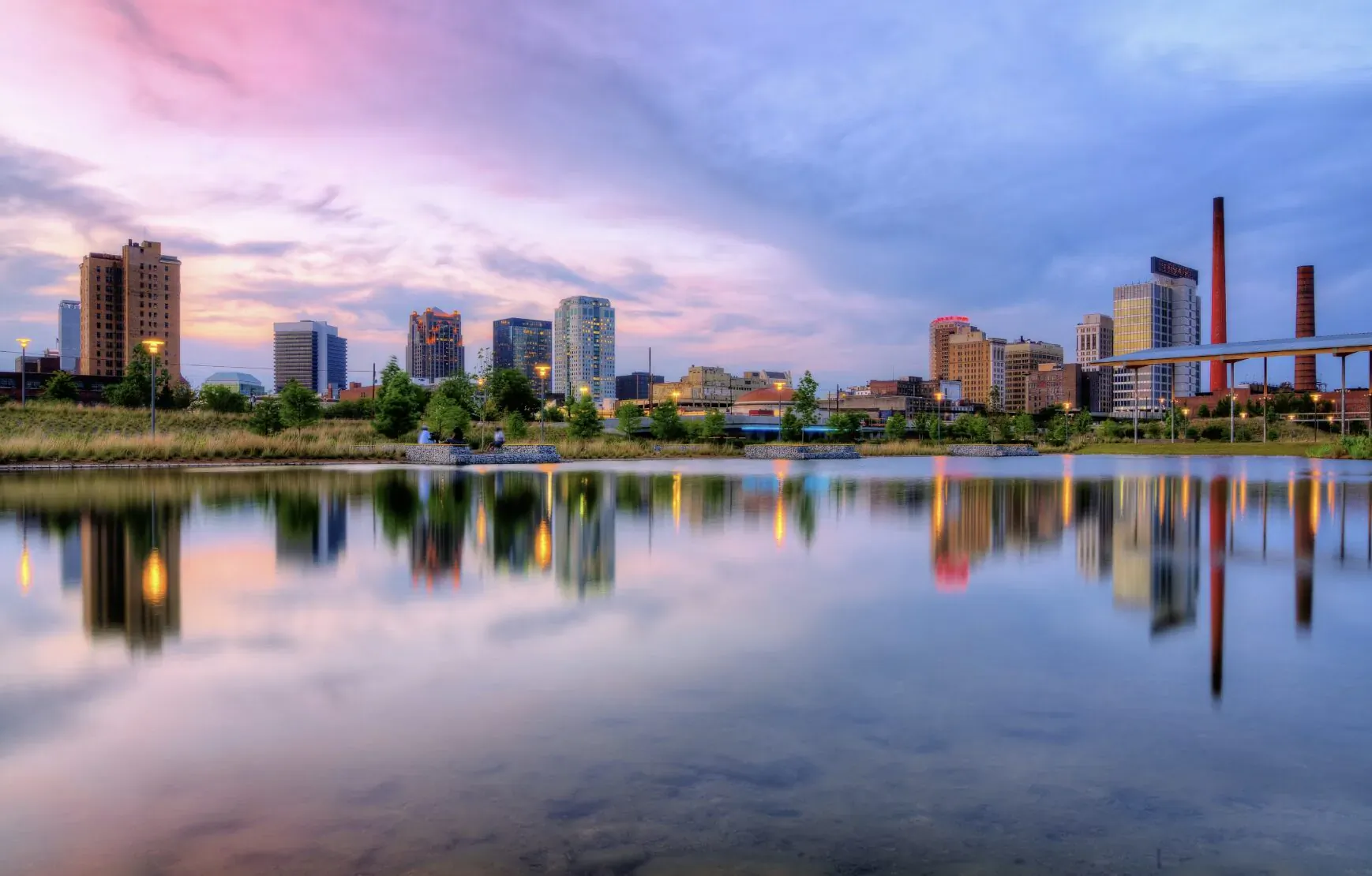 City skyline with high-rises, smokestacks, and bridge reflected in calm lake at pink sunset.