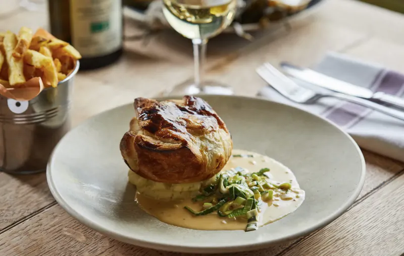 Puffed pastry pie on creamy sauce with greens in white bowl, fries in tin, white wine, on wooden table
