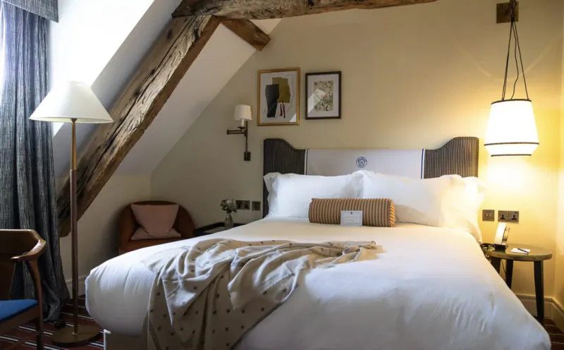 Cozy attic bedroom in Relais Henley with beamed ceiling, white bed with tan throw, lamp-lit, art on walls.
