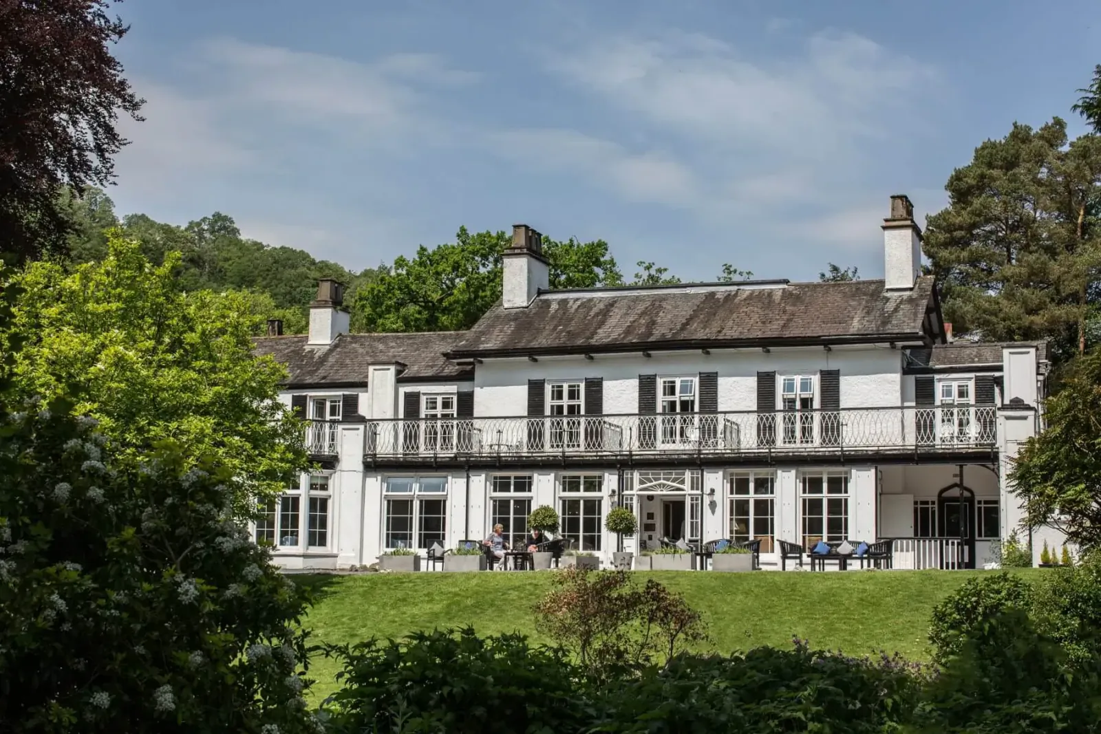 Rothay Manor Boutique Hotel, a white Georgian manor house with balcony, people at outdoor tables, surrounded by green hills and trees.